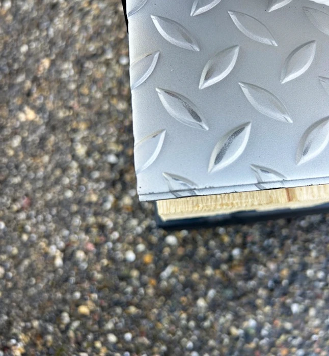 Coin d'une plaque de tôle larmée en aluminium gris posée sur un support en bois contreplaqué avec joint noir, photographiée en extérieur sur fond de béton désactivé granuleux